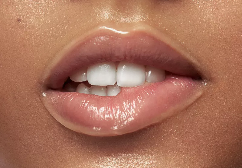 close up image of a woman's mouth, she is biting the edge of her lip. Her lips are glossy and pink